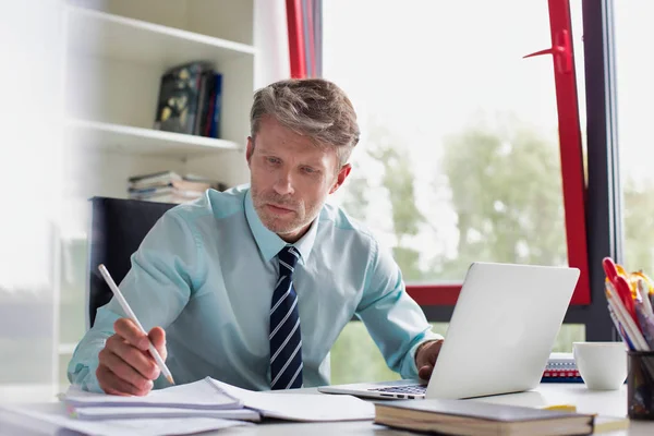 オフィスのデスクで書類事務をしながらノート パソコンを使用して深刻なビジネスマン — ストック写真