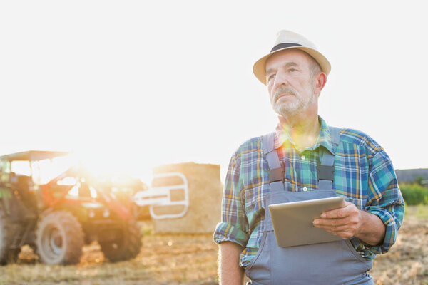 Portrait of confident senior farmer standing against tractor at farm with tablet