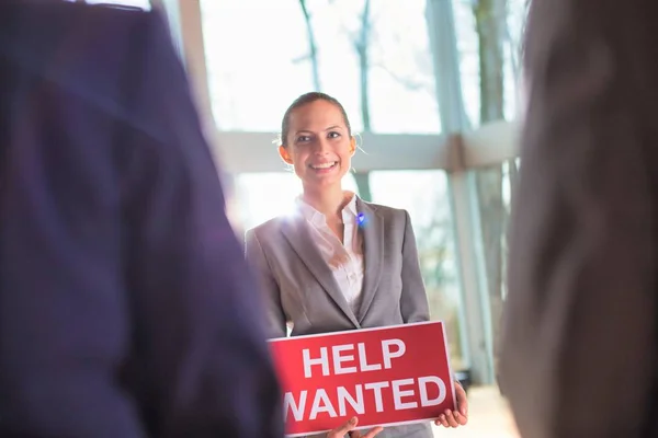 Confident young businesswoman holding red help wanted sign while
