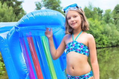 smiling girl in swimwear standing with pool raft at lakeshore