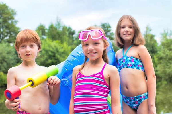 Portrait of smiling friends in swimwear standing with pool raft and squirt gun at lakeshore