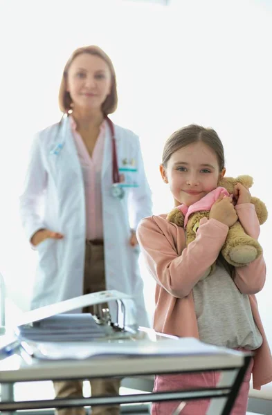 Ritratto di ragazza sorridente con orsacchiotto in piedi contro il medico — Foto Stock