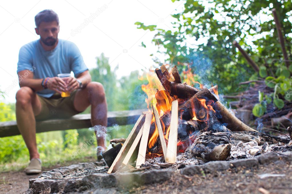 Man drinking beer while sitting on bench at park with bonfire