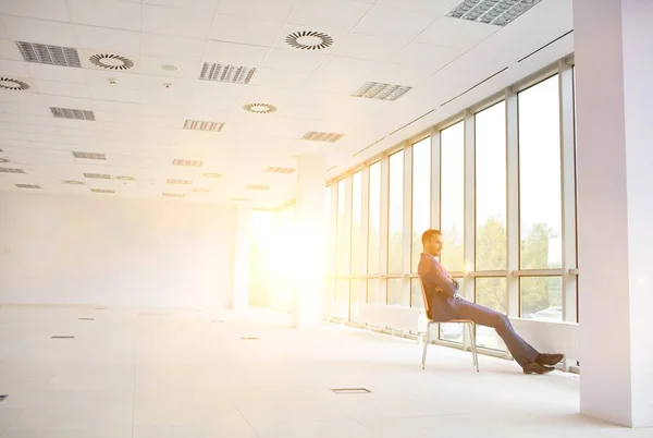 新しい空のオフィスの窓を見ながら椅子に座っている思慮深い若いビジネスマンの完全な長さ — ストック写真