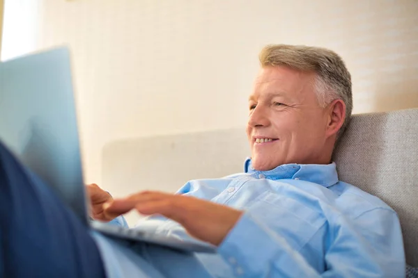 Χαμογελώντας ώριμος άνθρωπος χρησιμοποιώντας φορητό υπολογιστή, ενώ βρίσκεται στο κρεβάτι στο σπίτι — Φωτογραφία Αρχείου