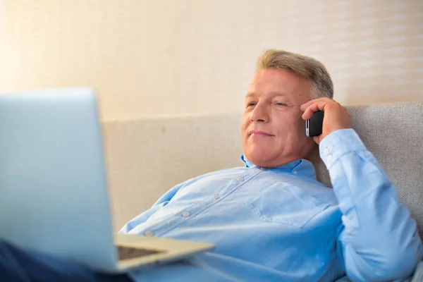 Lachende volwassen man met behulp van laptop tijdens het praten op de telefoon in be — Stockfoto