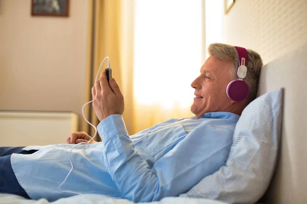 Mature man lying in bed while listening to music — Stock Photo, Image