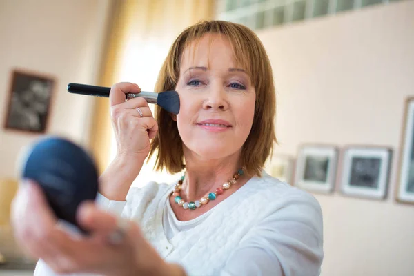Mujer madura aplicando maquillaje mientras está de pie en el dormitorio —  Fotos de Stock