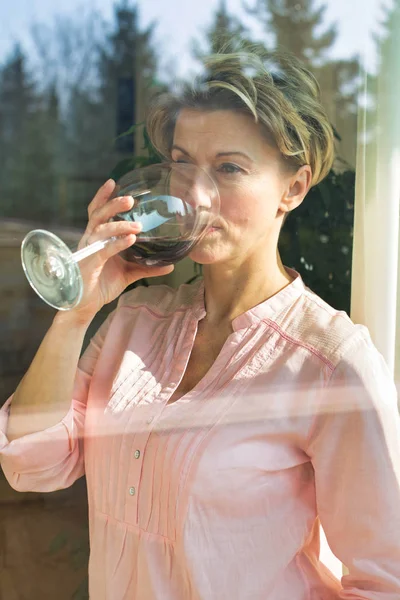 Sonriendo mujer madura sosteniendo wineglass visto a través de la ventana —  Fotos de Stock