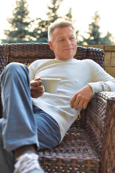 Usmívající se dospělý muž držící pití, zatímco sedí na proutěném křesle — Stock fotografie