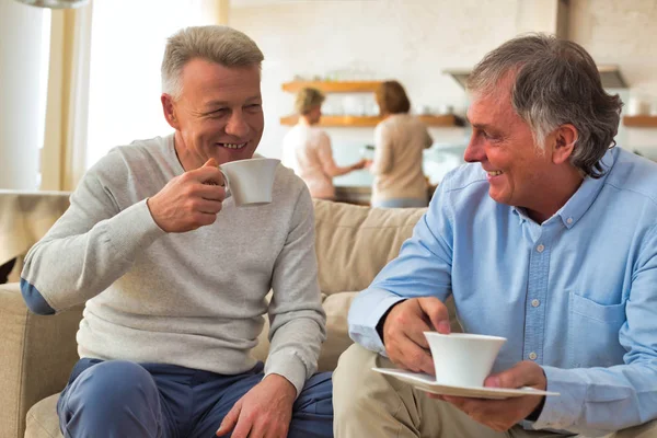 Улыбающиеся взрослые мужчины, держащие кофе, сидя дома на диване — стоковое фото