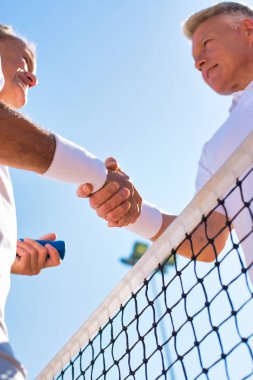 Erkekler açık gökyüzüne karşı tenis ağının başında el sıkışıyor.