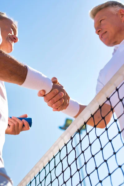 Men shaking hands while standing by tennis net against clear sky — ストック写真