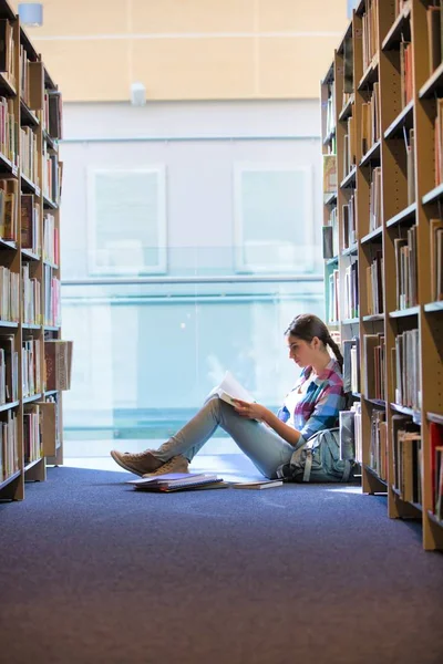 Student reading book while sitting against bookshelf at library — Stock Photo, Image