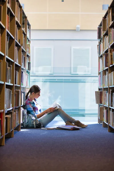 Studentin liest Buch, während sie in Bibliothek gegen Bücherregal sitzt — Stockfoto