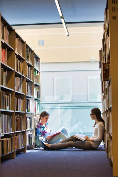 Estudiantes leyendo libro mientras están sentados contra estantería en la biblioteca —  Fotos de Stock