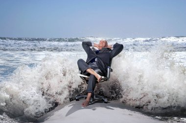 Senior business man sitting on office chair on beach being splashed by waves clipart