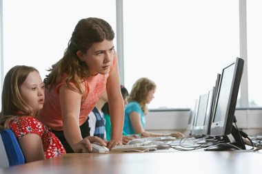 Bilgisayar laboratuvarında bilgisayar kullanan iki kız (10-12) arka planda çocuk