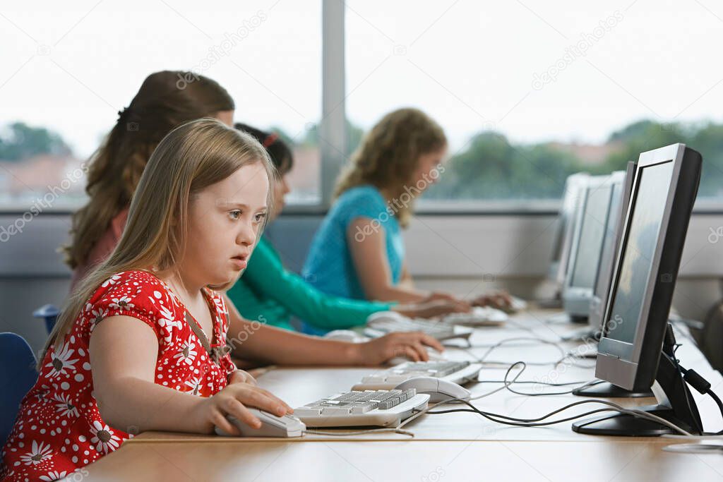 Girl (10-12) with Down syndrome using computer in computer lab children in background