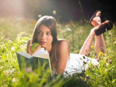 Genç bir kadın elinde kitap ve elmayla çayırda yatıyor.