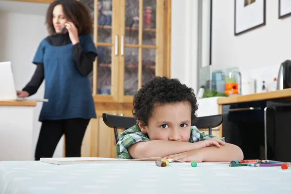 男の子の肖像 ラップトップと電話を使用してバックグラウンドでテーブルの母親に座って — ストック写真