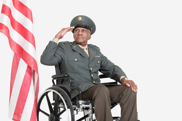 Senior male US military officer in wheelchair saluting American flag over gray background