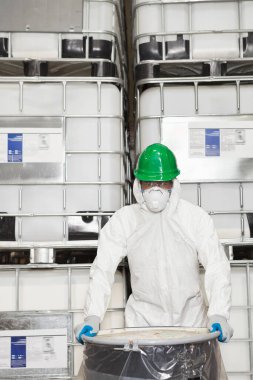 Tehlikeli madde giysisi giymiş teknisyen elinde plastik kaplı kimyasal varil tutuyor.