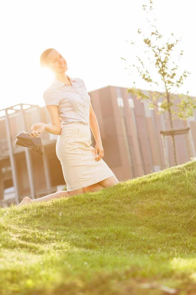 Uśmiechnięta Młoda Bizneswoman Trzymająca Wysokie Obcasy Klęcząc Trawniku Biurowym — Zdjęcie stockowe