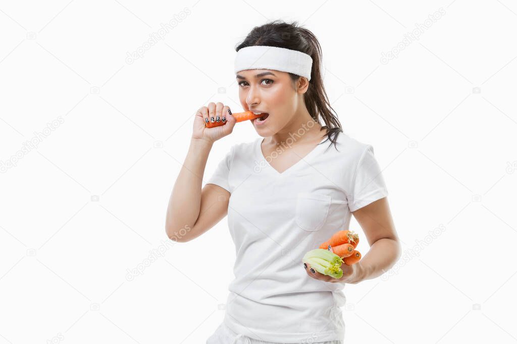 Portrait of young sportswoman eating carrot over white background