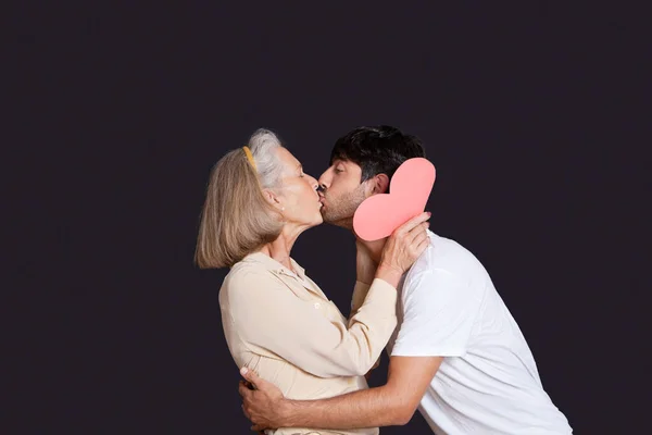 Jovem Beijando Mulher Sênior Com Coração Papel Vermelho Contra Fundo — Fotografia de Stock
