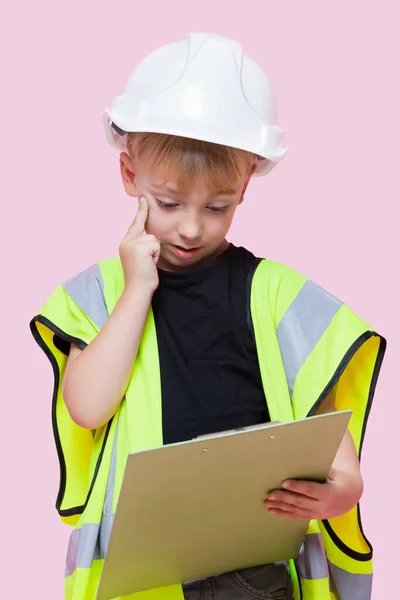 一个体贴入微的小男孩打扮成建筑工人 用粉红背景看剪贴板的画像 — 图库照片