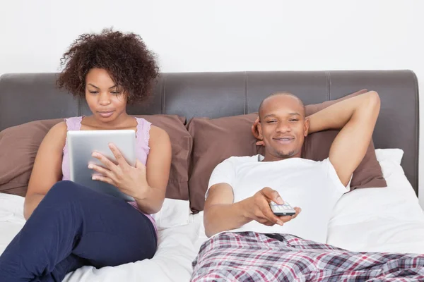 Νεαρός Άνδρας Βλέποντας Τηλεόραση Ενώ Γυναίκα Χρησιμοποιεί Tablet Στο Κρεβάτι — Φωτογραφία Αρχείου