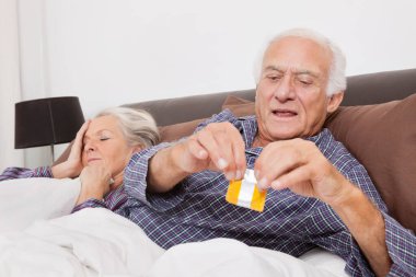 Kocası yatakta uyurken prezervatif paketini yırtan yaşlı bir adam.