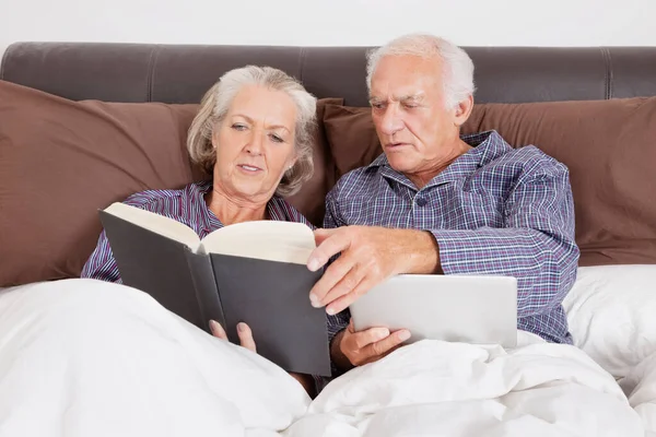 部屋にベッドの上に横たわっている間 デジタルタブレット読書本を持つシニアカップル — ストック写真