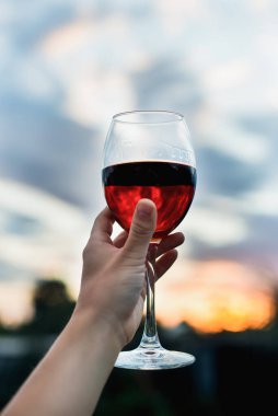 Bir kadının eli, günbatımı gökyüzünün arka planında bir kadeh kırmızı şarap tutar.