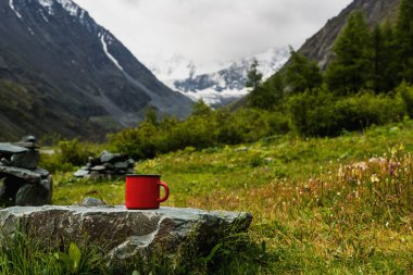 Çay ya da kahveli kırmızı bir kupa kar kaplı Altai dağlarının arka planında bir taşın üzerinde duruyor.