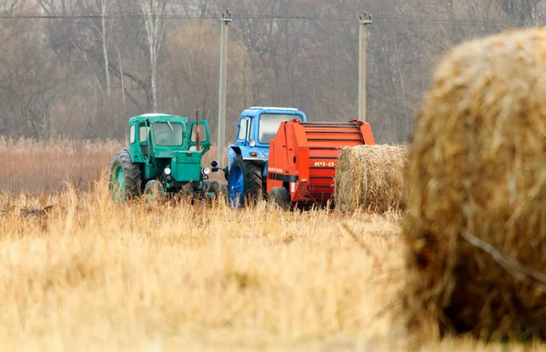 Ciągniki do zbioru suchej trawy w bele słomy w polu jesienią. Specjalne maszyny rolnicze. Na pierwszym planie są bele słomy. Płytkie głębia ostrości. — Zdjęcie stockowe