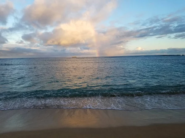Rainbow shines through clouds over ocean off Kahala Beach with boat on the horizon on Oahu, Hawaii.
