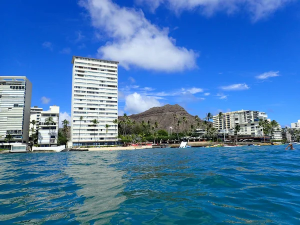 Hotel building, Outrigger Canoe Club, coconut trees, Condo buildings, clouds, and Diamond Head Crater in the distance on Oahu, Hawaii viewed from the water on a beautiful day.