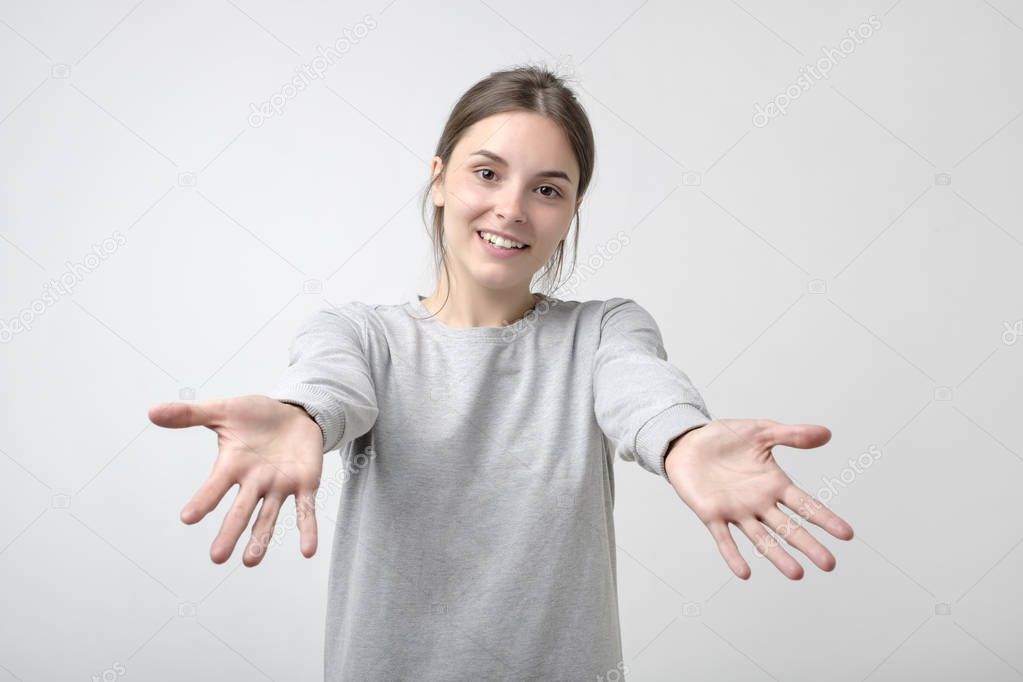 portrait of attractive smiling teenage girl raised up arms hands at you. She is dressed in gray simple t-shirt. Happy to welcome a friend or guest