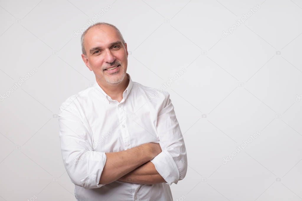 Confident senior man in white t-shirt crossing hands on chest and looking at camera while standing against gray background. Self confident hispanic senior.