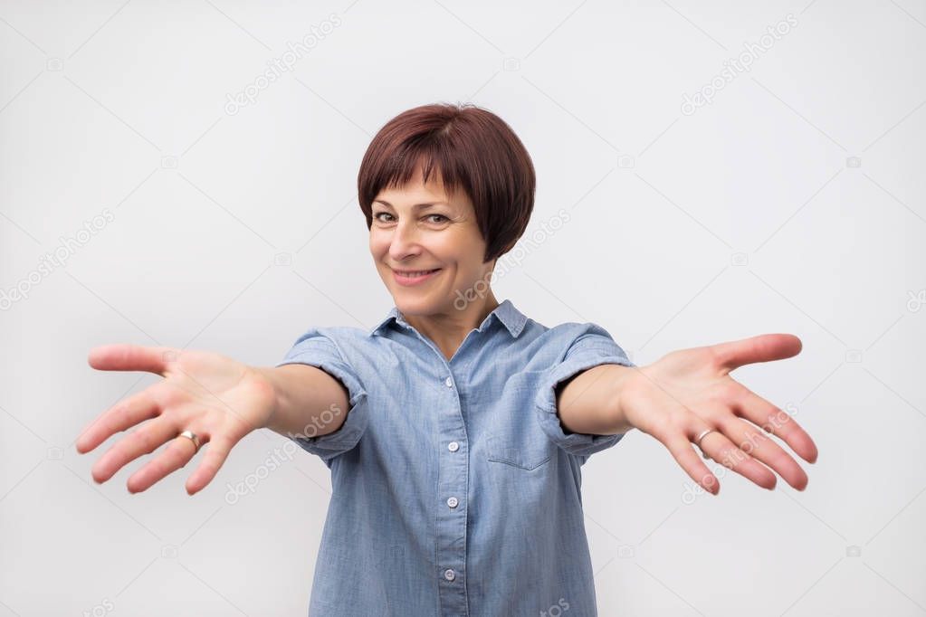 Portrait of attractive mature woman raised up arms hands at you. She is dressed in blue shirt.