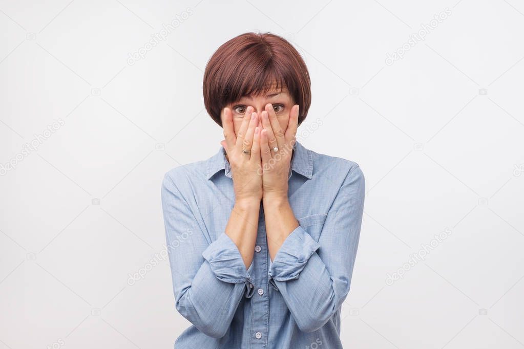 Close up portrait of attractive, mature woman in blue shirt closing her face with fingers. She is worried.