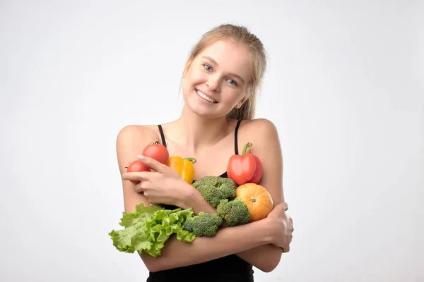 Mujer rubia sonriente sosteniendo una pila de verduras frescas saludables de pie sobre fondo gris — Foto de Stock