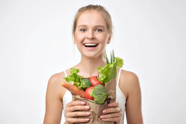 灰色の背景の上に立っての新鮮な野菜の山を保持している金髪の若い女性の笑みを浮かべてください 健康的な食事を食べることの概念 — ストック写真