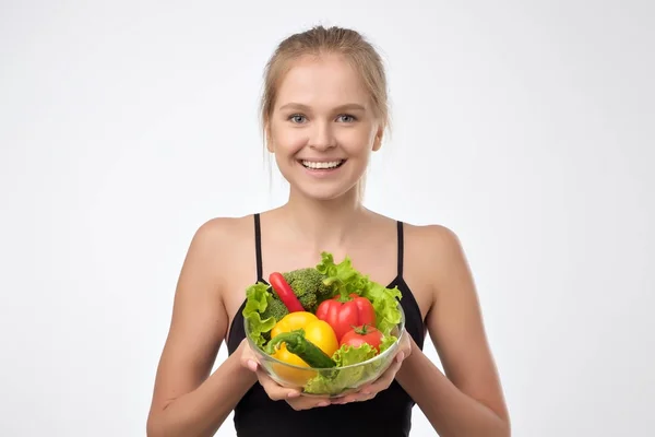 Sorrindo loira jovem segurando uma pilha de legumes frescos saudáveis em pé sobre fundo branco . — Fotografia de Stock