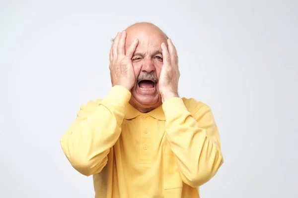 Старший іспаномовний чоловік в жовтій сорочці тягне шкіру на обличчя руками. Він боїться або втомився від стресу . — стокове фото