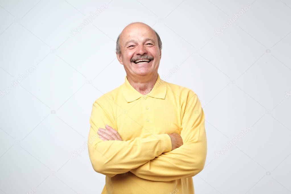 Charming handsome senior man in casual clothes keeping arms crossed and laughing while standing isolated on white background.