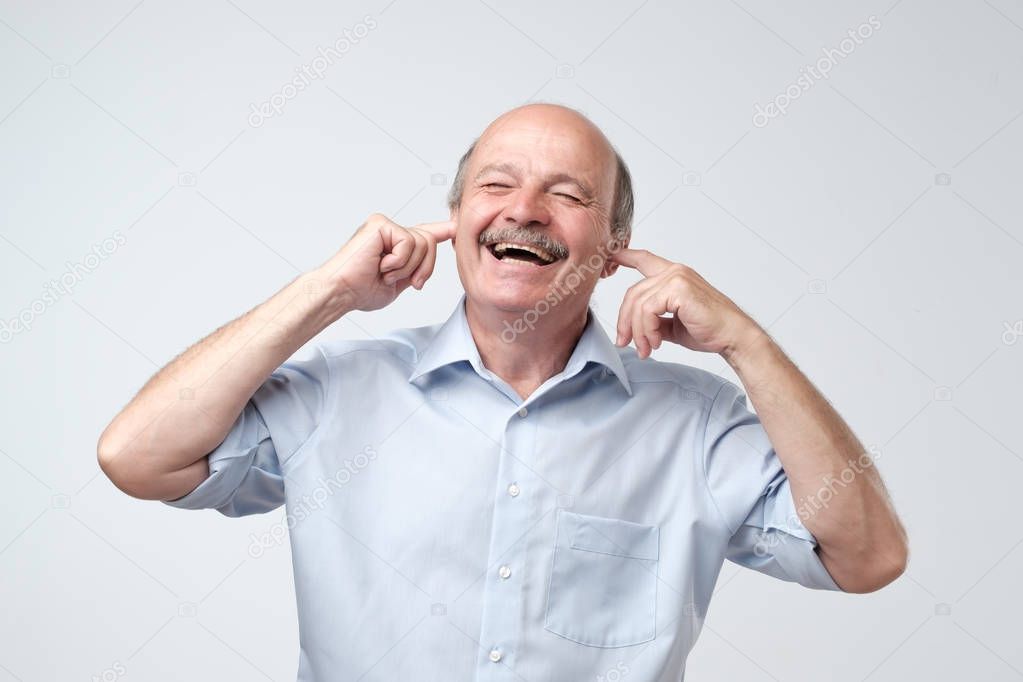 Using natural earplugs not to hear spoilers. Good-looking happy mature european man smiling with closed eyes, holding index fingers in ears, waiting for surprise