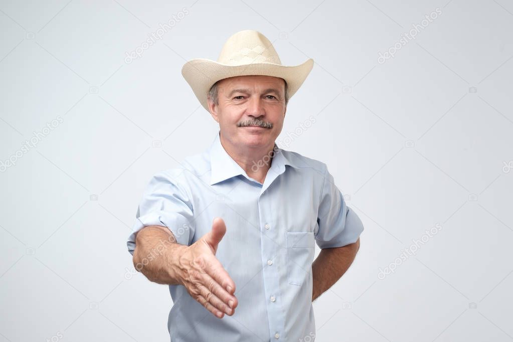 You are welcome concept. Cheerful mature man in blue shirt and cowboy hat gesturing welcome sign and smiling while standing against gray wall. He is happy to meet dear guests